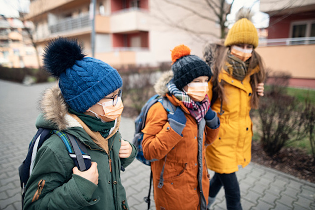 Children in winter coats and hats wearing face masks and walking on sidewalk.  