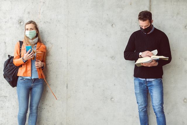 A woman student wearing a face mask looks at her phone while a few feet away a male student wearing a face mask looks at a book. 