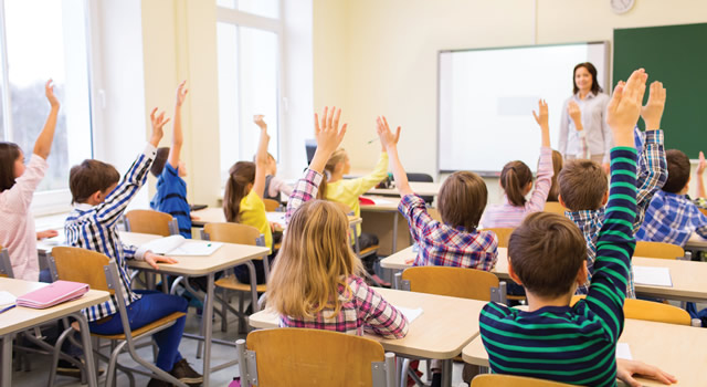 a group of studenst in a classroom all raising hands