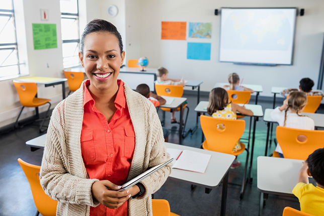 A teacher carrying a tablet smiles while students are working at their desks facing the front of the classroom.