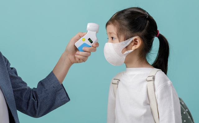 Woman taking the temperature of a child who is wearing a face mask and a backpack with a contactless thermometer. 